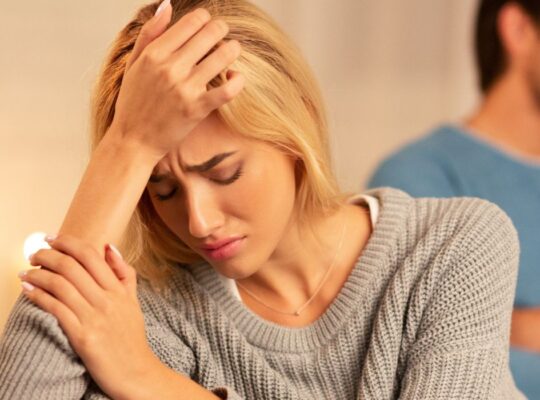 Image of a woman upset after discovery of her partners betrayal. He sits in the background looking moody and ignoring her pain.
